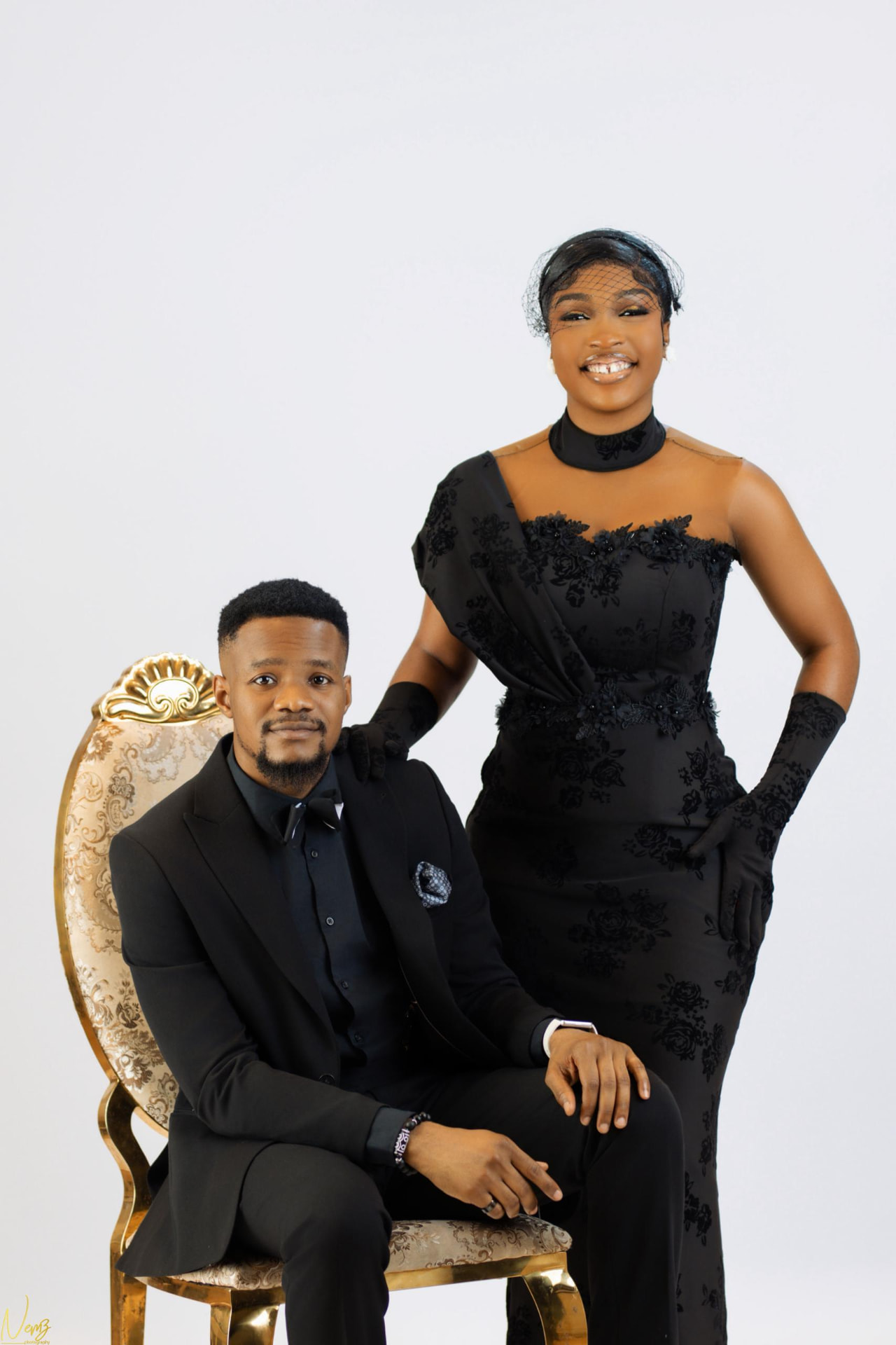 Iheoma in a black lace gown standing beside Kent seated in a gold armchair